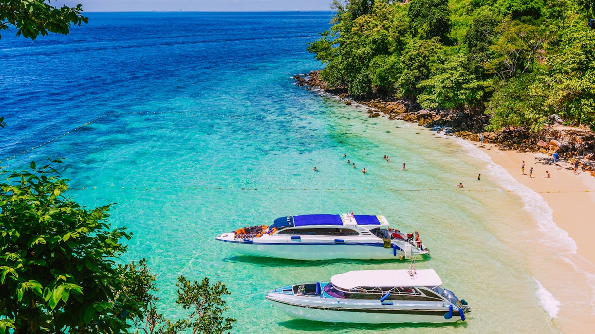 Ferry boats docked on a tropical beach in Phuket, Thailand, with clear blue water.