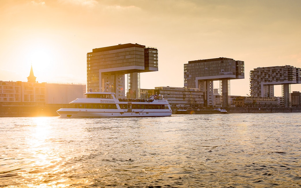 Cruise boat on the Rhine River with Cologne's Kranhäuser buildings at sunset.