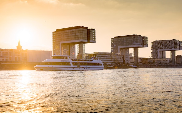 Cruise boat on the Rhine River with Cologne's Kranhäuser buildings at sunset.