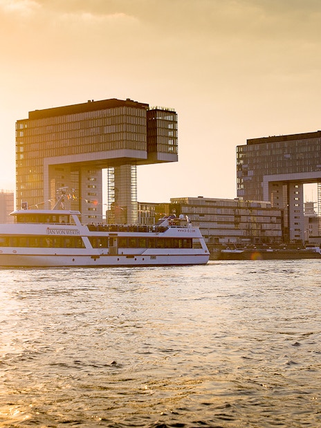 Cruise boat on the Rhine River with Cologne's Kranhäuser buildings at sunset.