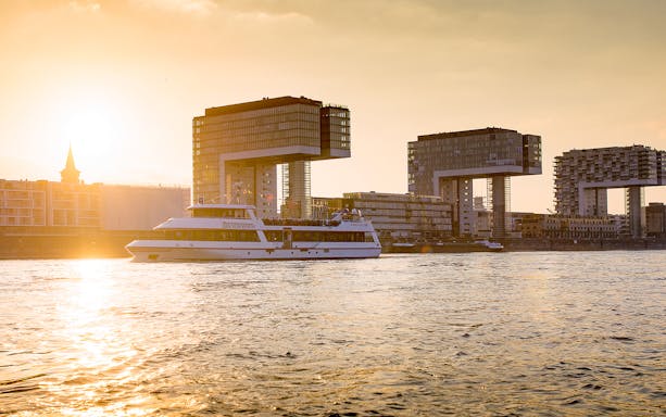 Cruise boat on the Rhine River with Cologne's Kranhäuser buildings at sunset.