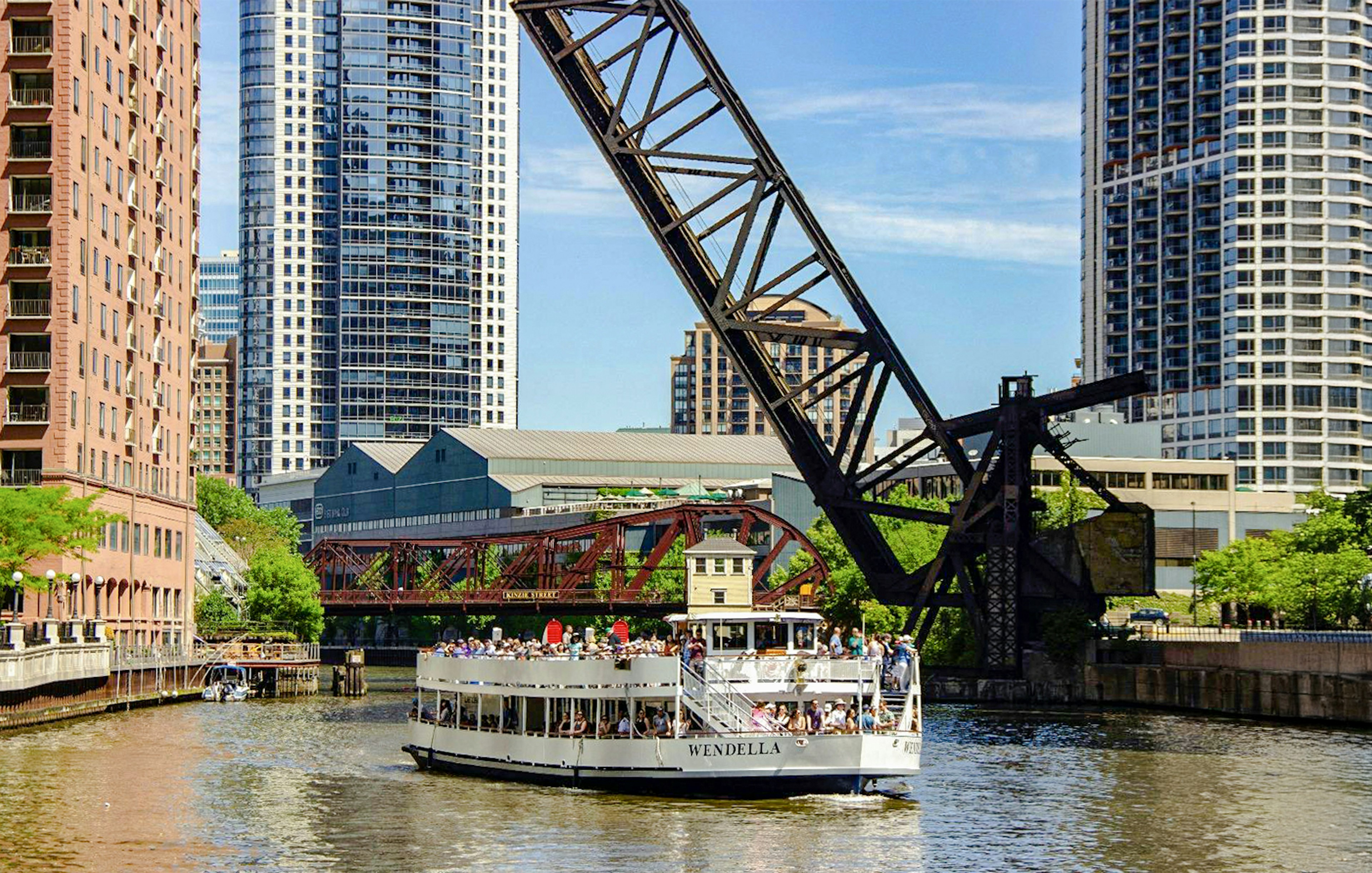 Chicago River architecture cruise boat passing under a lift bridge with city skyscrapers in the background.