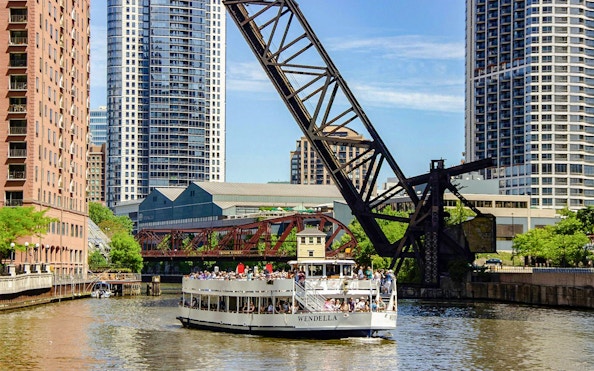 Chicago River architecture cruise boat passing under a lift bridge with city skyscrapers in the background.