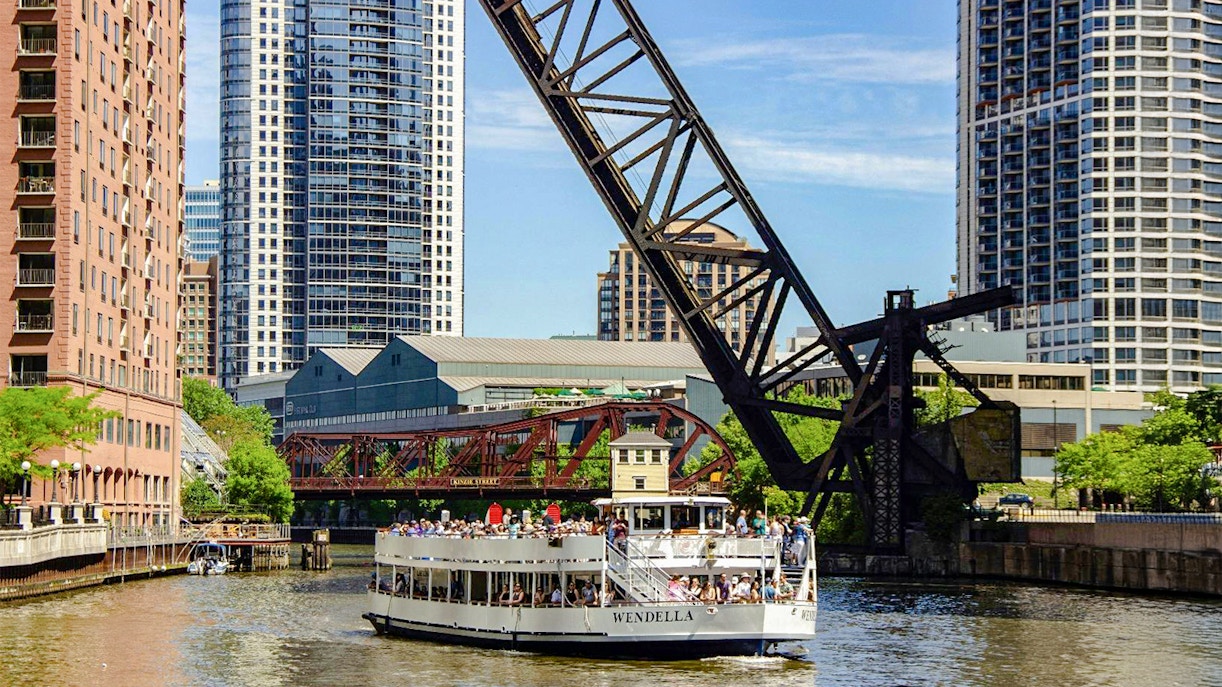 Chicago River architecture cruise boat passing under a lift bridge with city skyscrapers in the background.