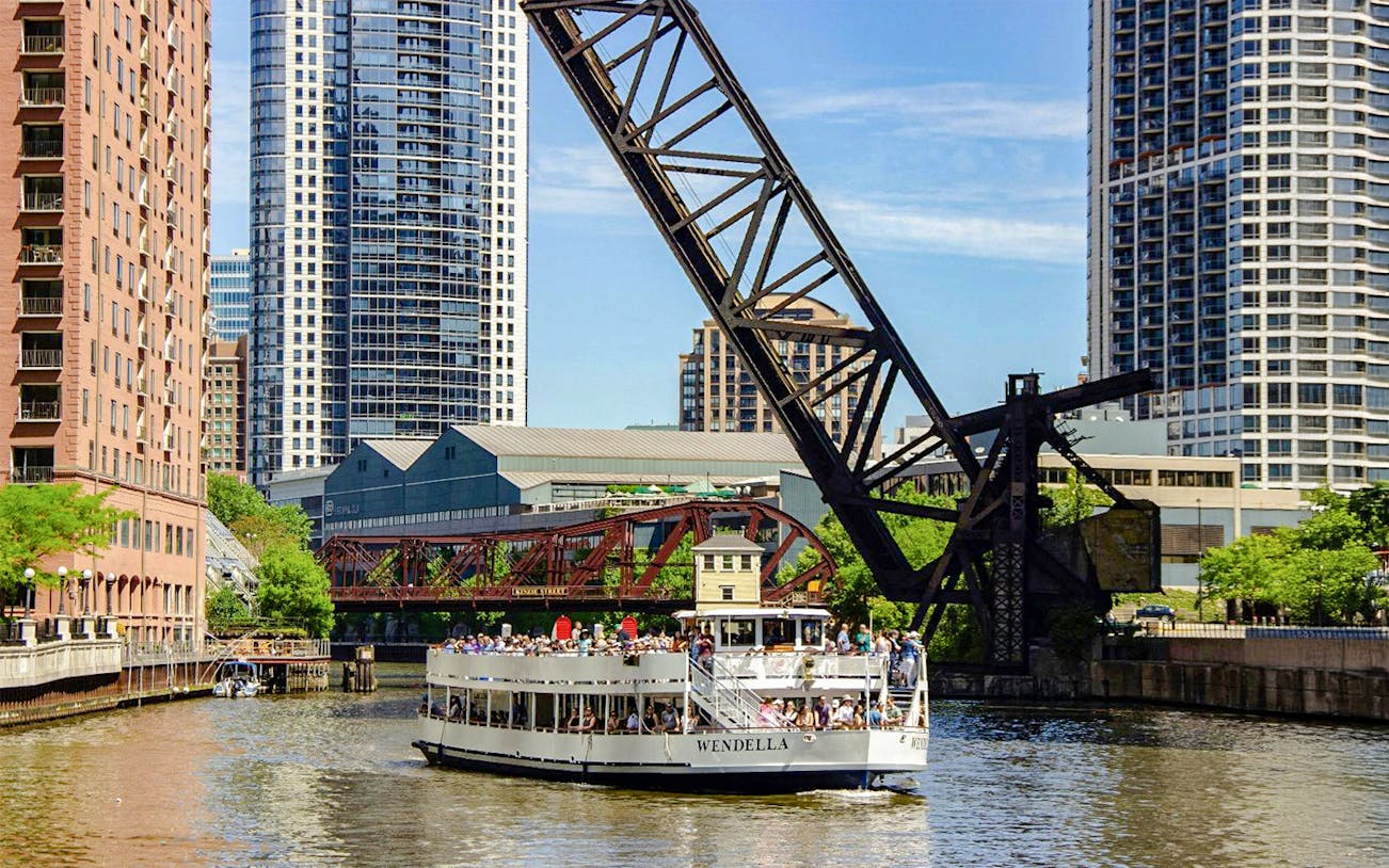 Chicago River architecture cruise boat passing under a lift bridge with city skyscrapers in the background.