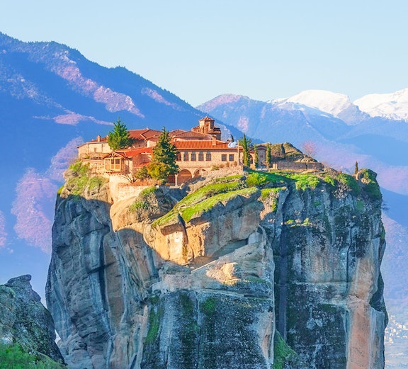 Monastery atop rock formation in Meteora, Greece, with mountains in the background.