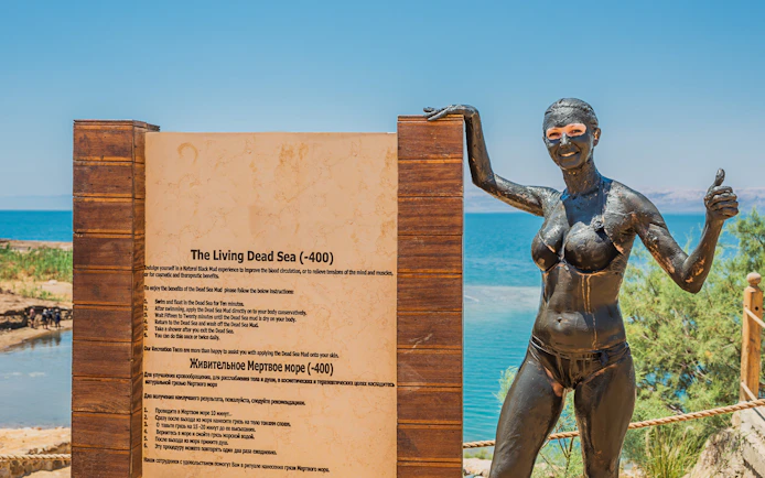 Tourist covered in Dead Sea mud standing by informational sign at the Dead Sea.