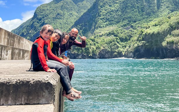 Family in wetsuits sitting by the ocean, ready for a surf lesson in a scenic coastal area.