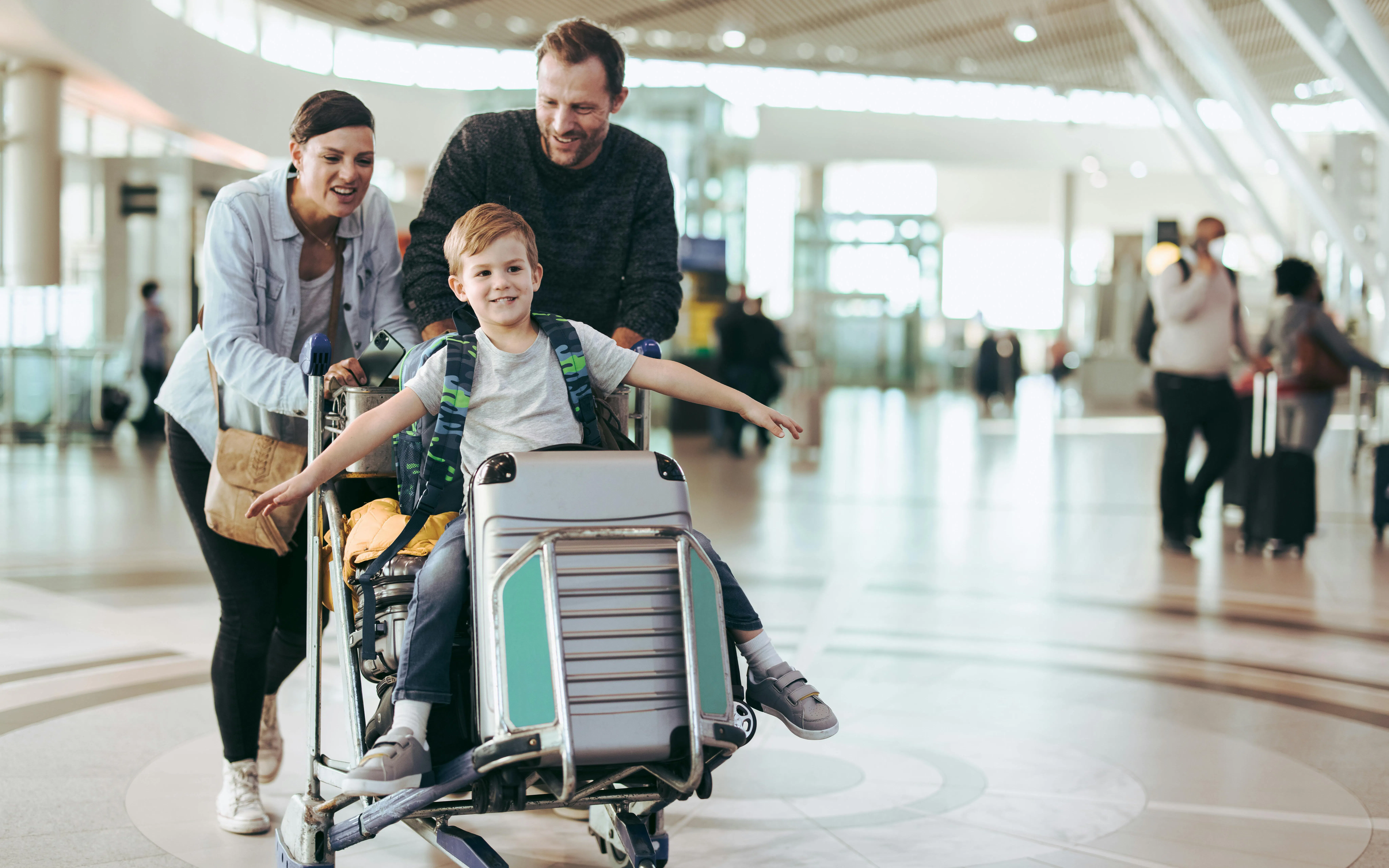 Parents pushing luggage trolley with son at airport terminal.