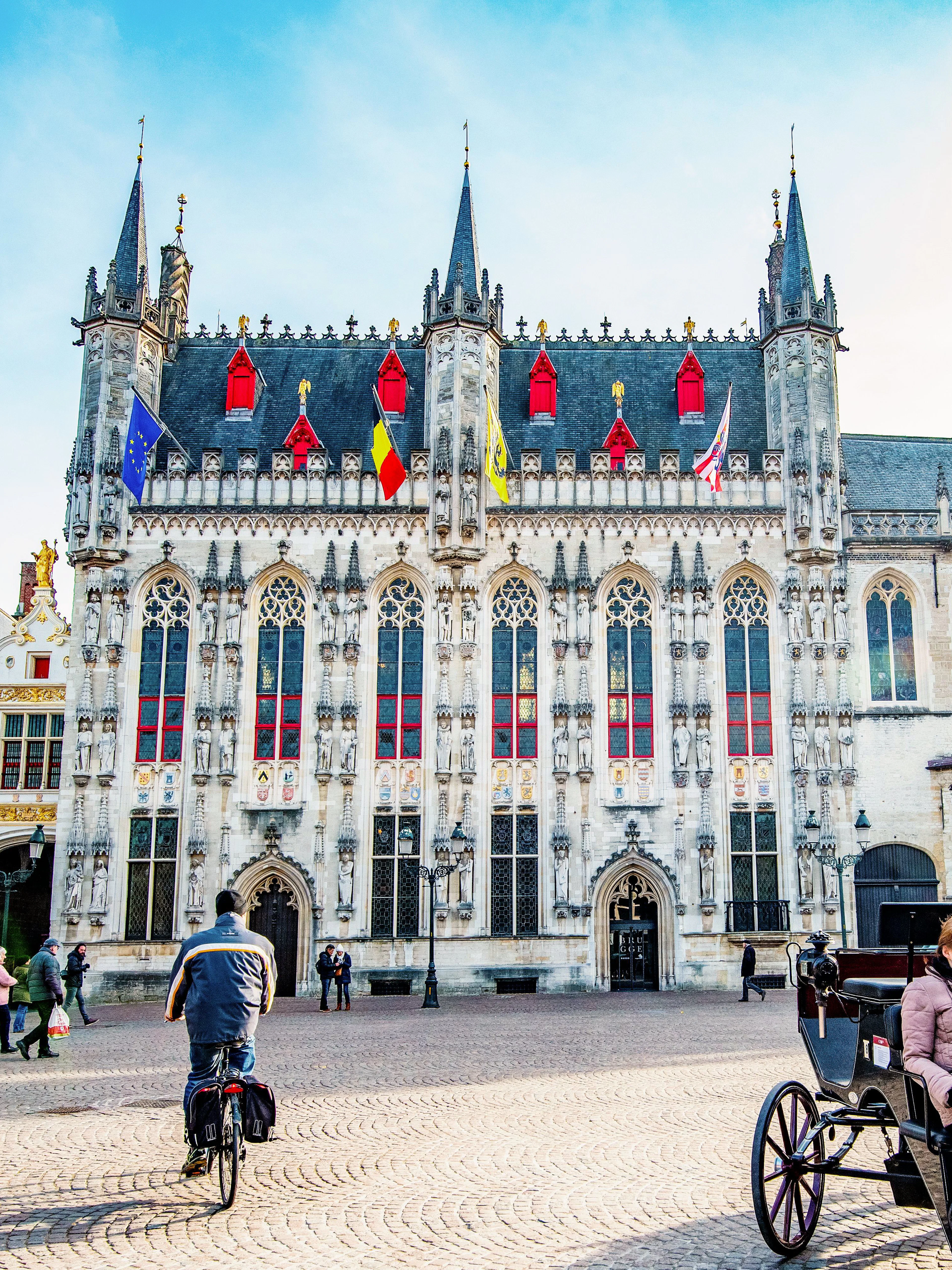 City Hall in Bruges with people cycling and a horse-drawn carriage in the square.