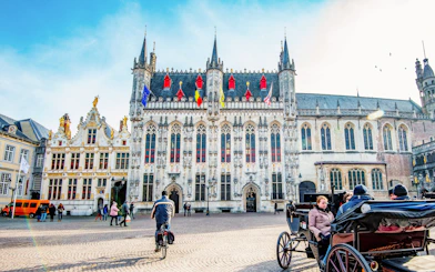 City Hall in Bruges with people cycling and a horse-drawn carriage in the square.