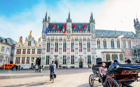 City Hall in Bruges with people cycling and a horse-drawn carriage in the square.