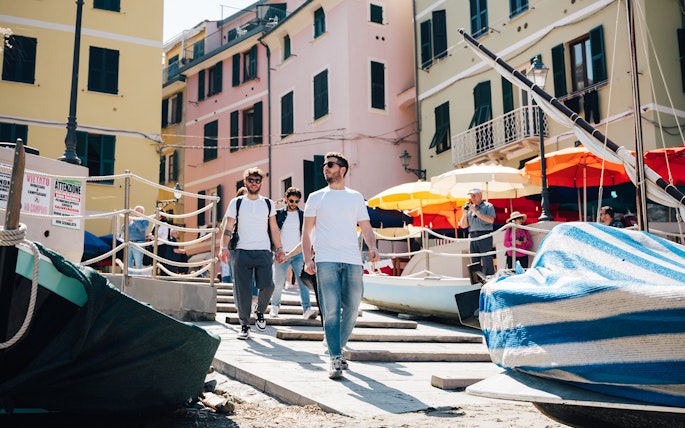Tourists walking through colorful streets of Cinque Terre, Italy, with boats and umbrellas nearby.