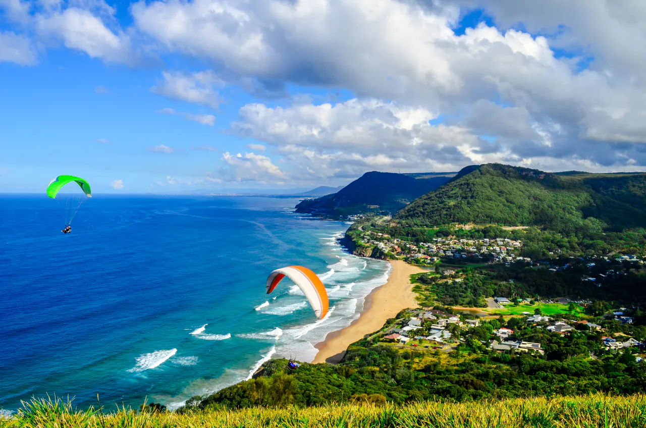Paragliders over coastal landscape near Sydney, showcasing beach and lush hills.