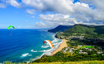 Paragliders over coastal landscape near Sydney, showcasing beach and lush hills.
