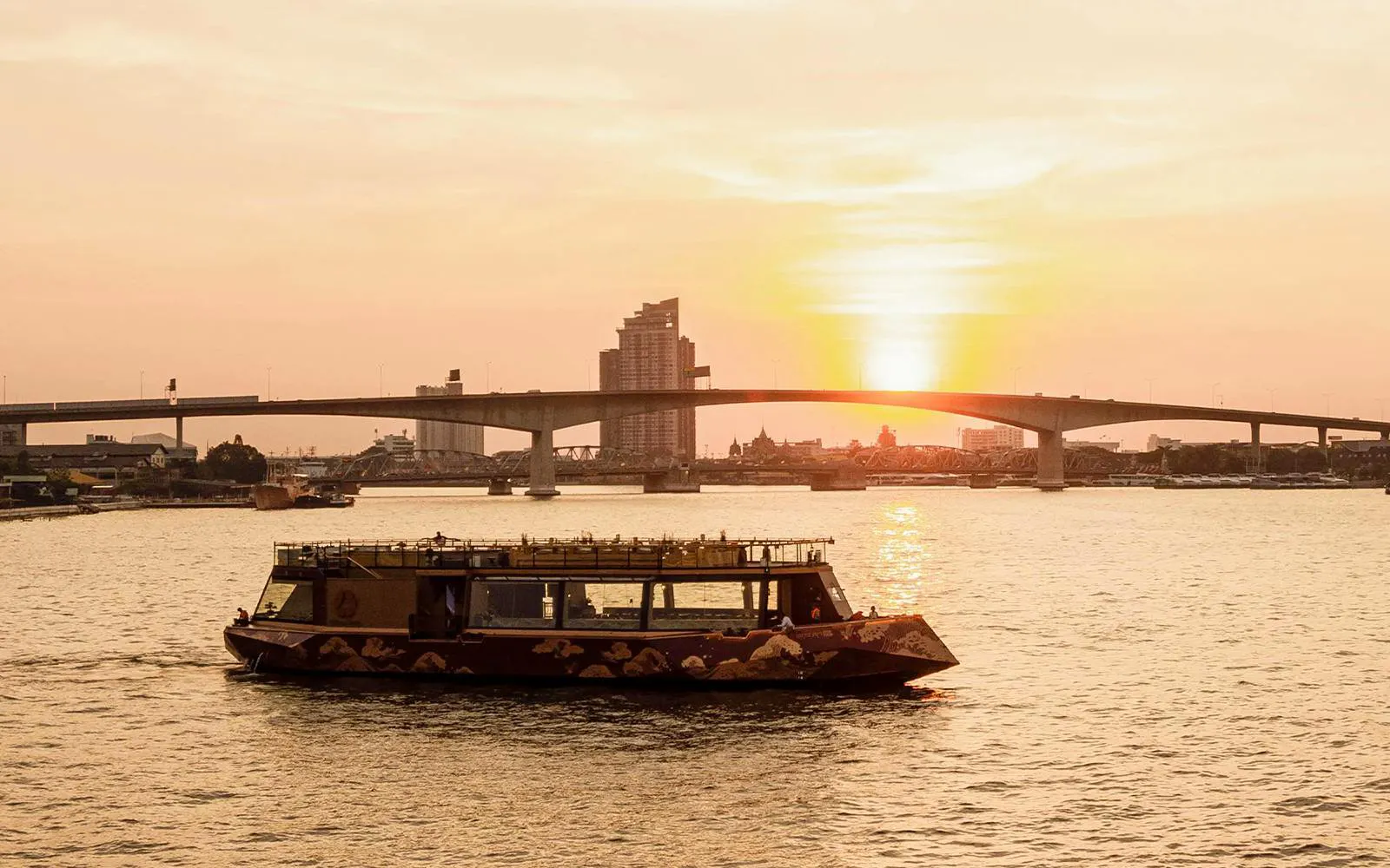 Cruise boat on Chao Phraya River at sunset, Bangkok bridge in background.
