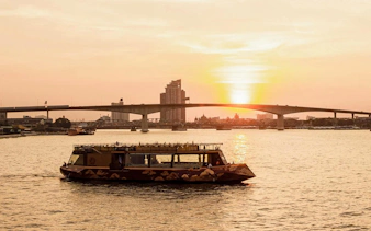 Cruise boat on Chao Phraya River at sunset, Bangkok bridge in background.