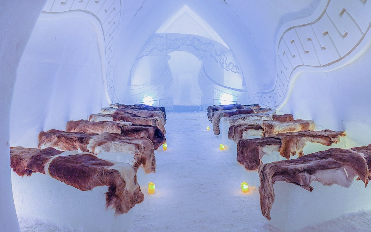 Inside view of Arctic SnowHotel with ice benches covered in fur and ambient lighting.