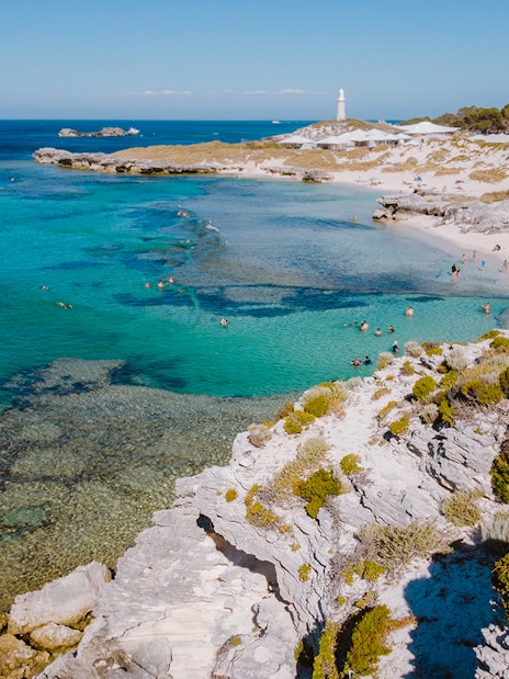 Tourists swimming at a beach on Rottnest Island with a lighthouse in the distance.