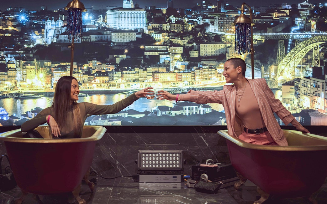 Two people in bathtubs toasting with drinks at Pink Palace, cityscape backdrop.