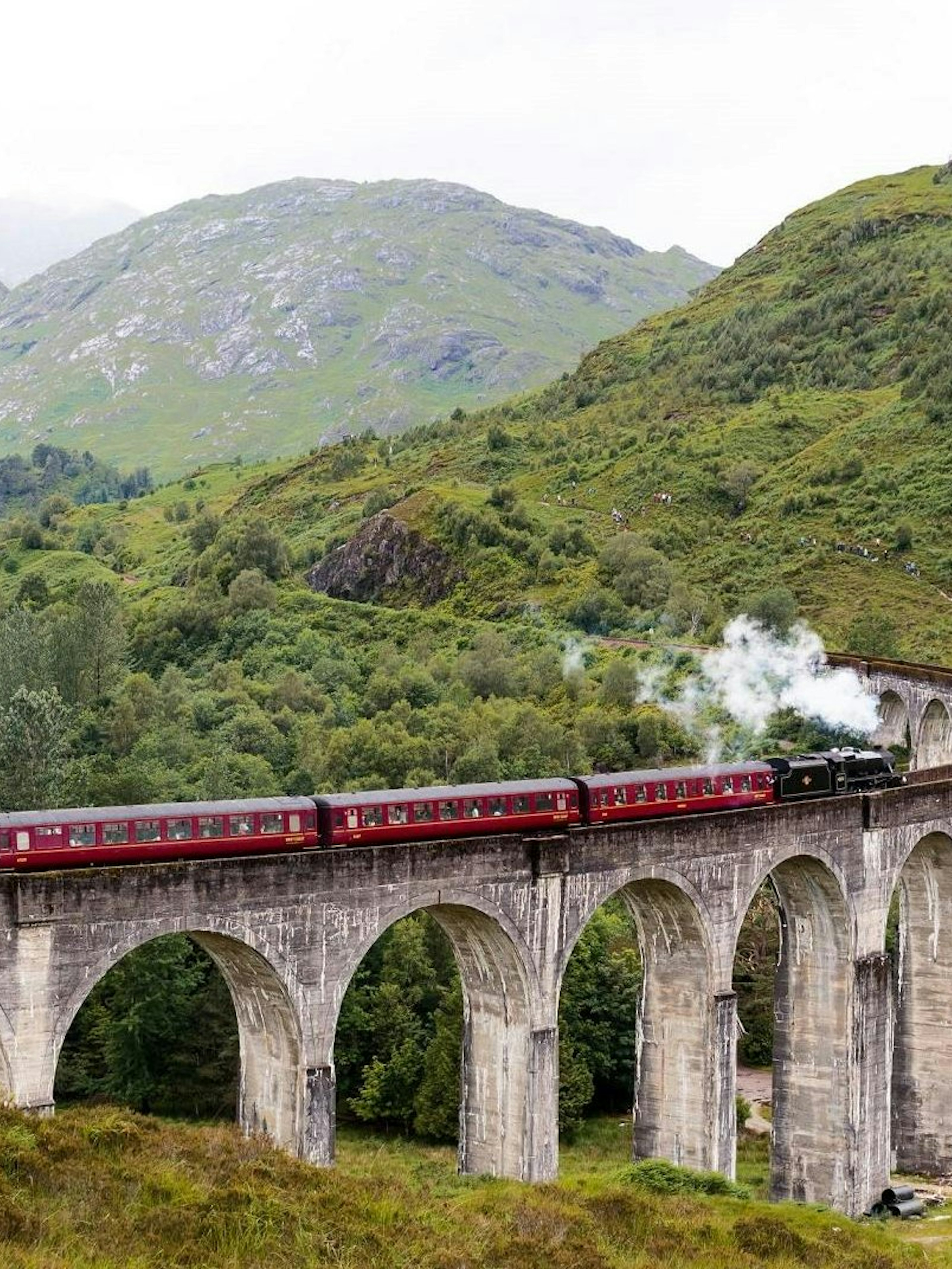 Steam train crossing the Glenfinnan Viaduct in Scotland with lush green hills in the background.