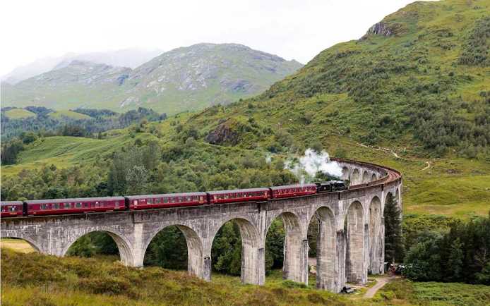 Steam train crossing the Glenfinnan Viaduct in Scotland with lush green hills in the background.