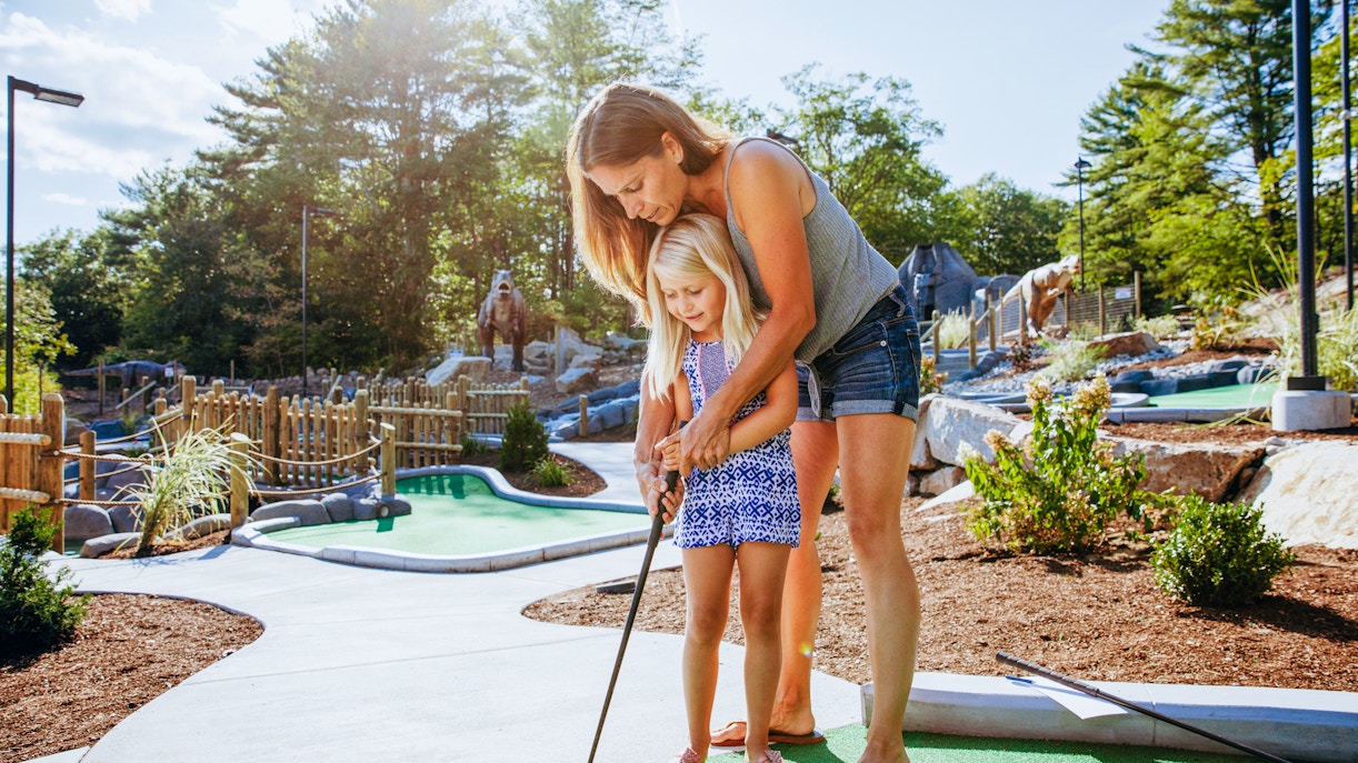 Woman guiding daughter at a miniature golf course with dinosaur statues in the background.