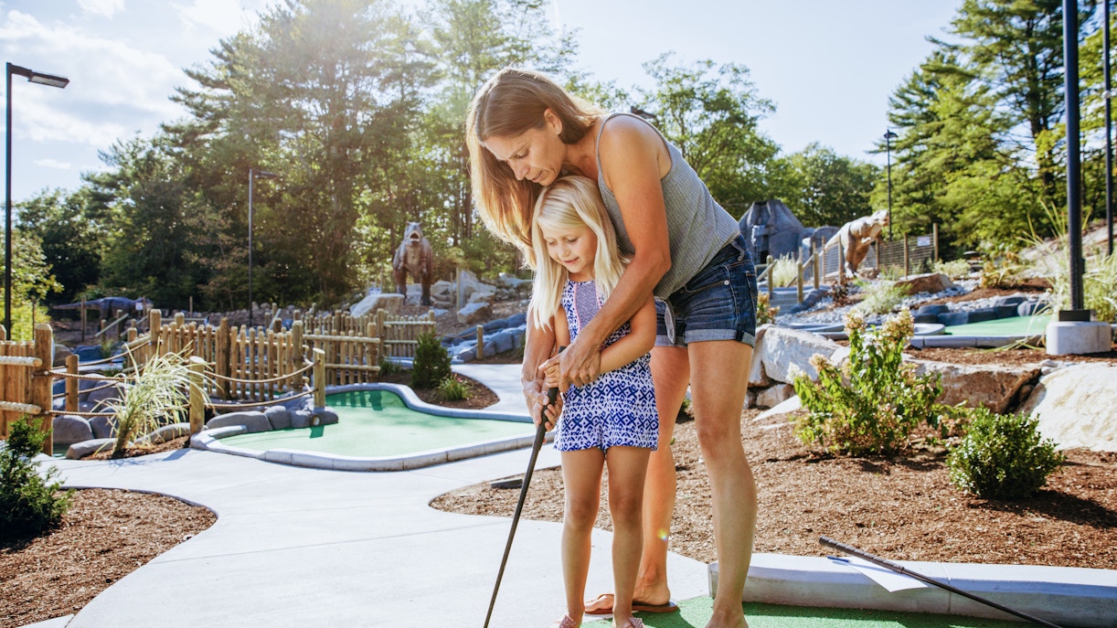 Woman guiding daughter at a miniature golf course with dinosaur statues in the background.