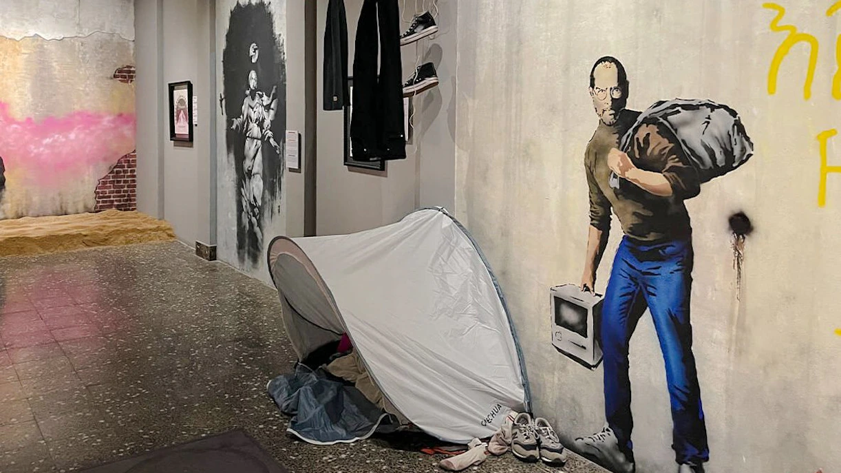 Artwork display at Banksy Museum Amsterdam featuring a mural of a man with a sack and a tent installation.
