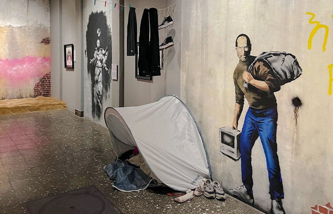 Artwork display at Banksy Museum Amsterdam featuring a mural of a man with a sack and a tent installation.