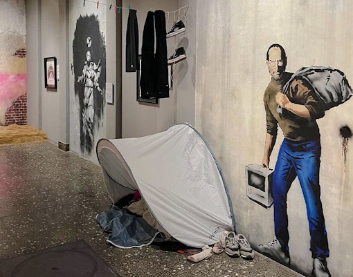 Artwork display at Banksy Museum Amsterdam featuring a mural of a man with a sack and a tent installation.