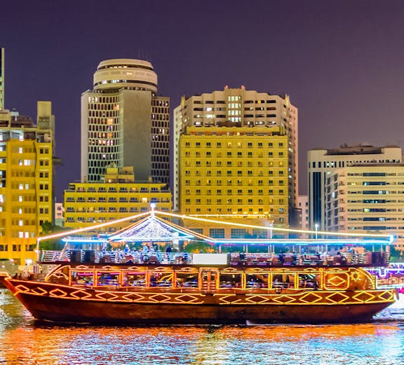 Traditional dhow boat illuminated on Dubai Creek with city skyline in the background.