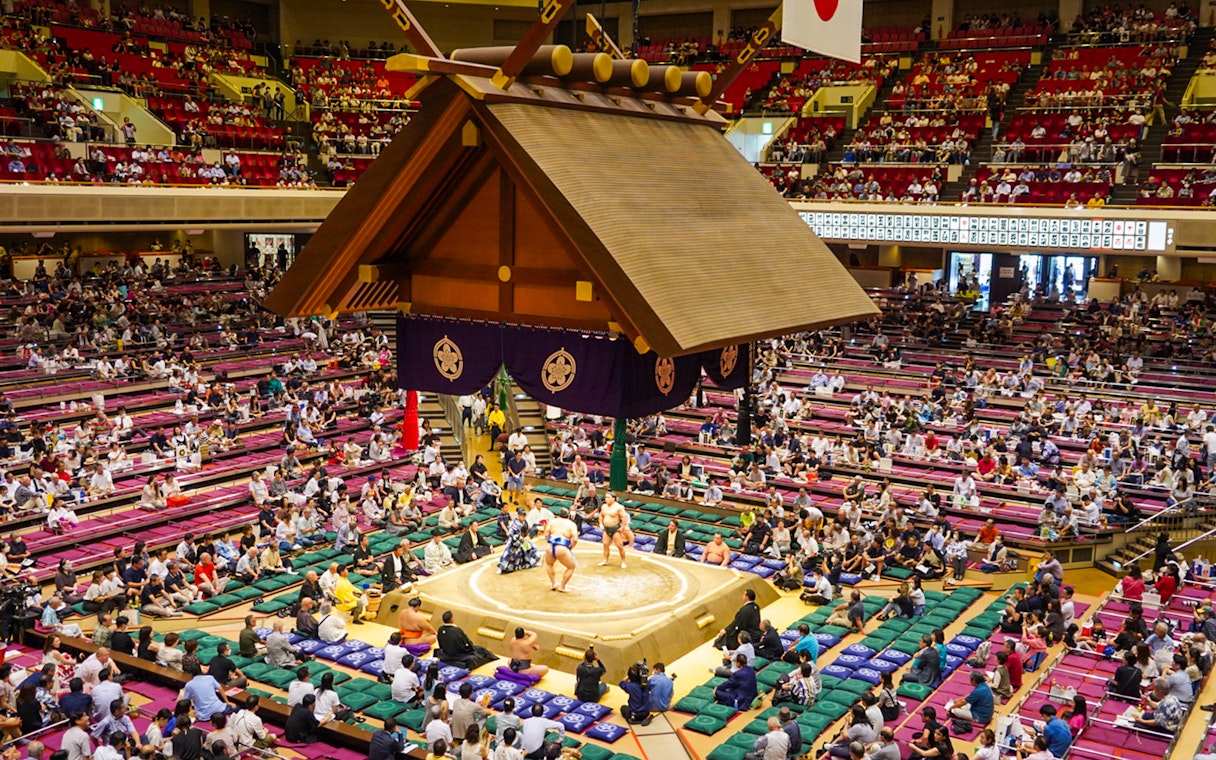 Sumo wrestlers competing in Ryogoku arena, Tokyo, with audience in tiered seating.