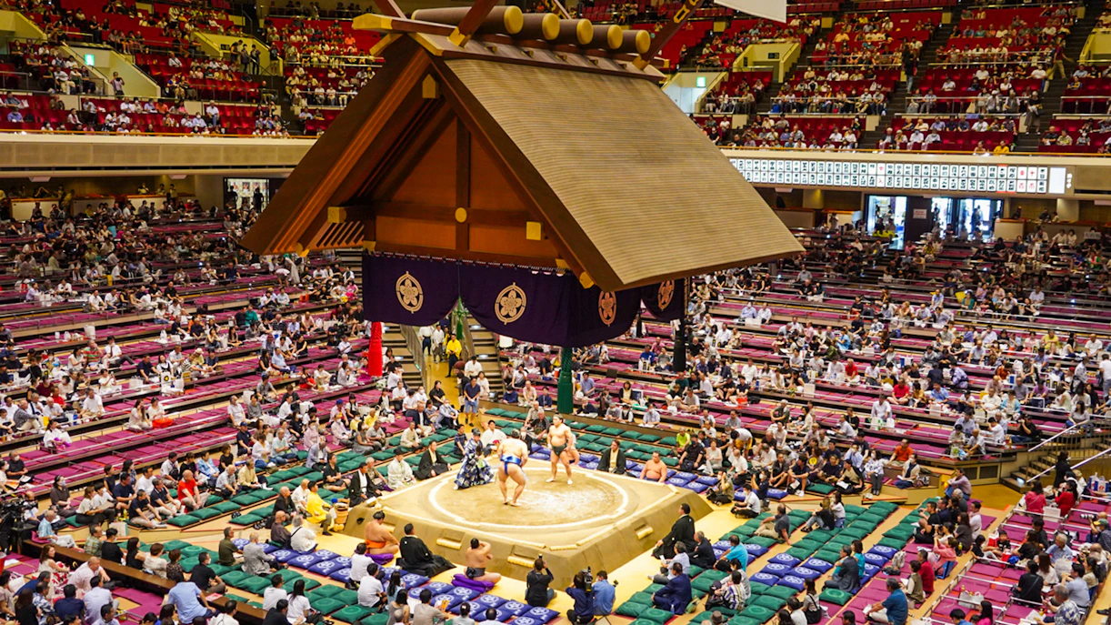 Sumo wrestlers competing in Ryogoku arena, Tokyo, with audience in tiered seating.