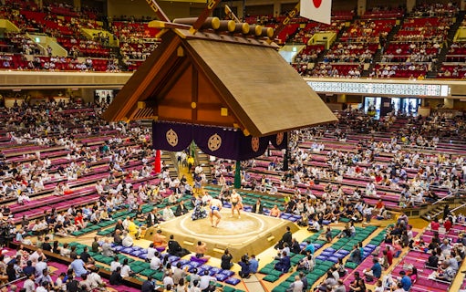 Sumo wrestlers competing in Ryogoku arena, Tokyo, with audience in tiered seating.