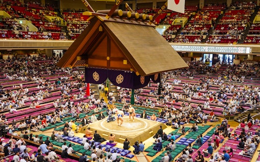 Sumo wrestlers competing in Ryogoku arena, Tokyo, with audience in tiered seating.