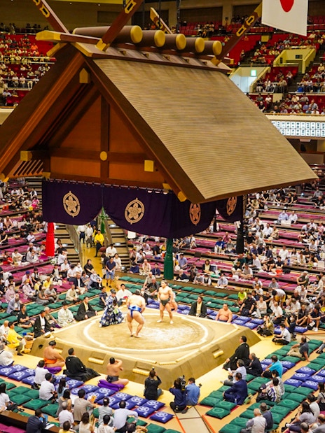 Sumo wrestlers competing in Ryogoku arena, Tokyo, with audience in tiered seating.