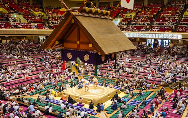 Sumo wrestlers competing in Ryogoku arena, Tokyo, with audience in tiered seating.