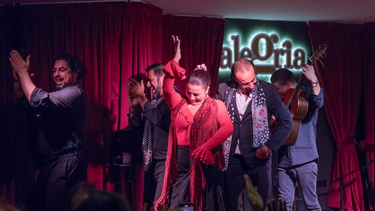 Dançarina de flamenco mlaga em movimento, vestindo um vestido branco com babados, sob luzes quentes no palco.