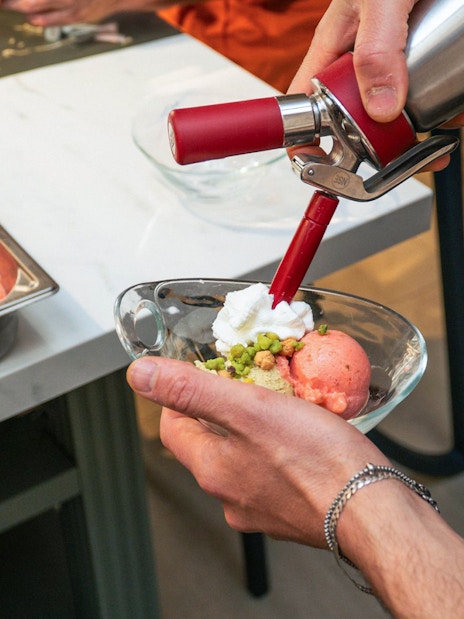 Person adding whipped cream to gelato in a dish with pistachios.