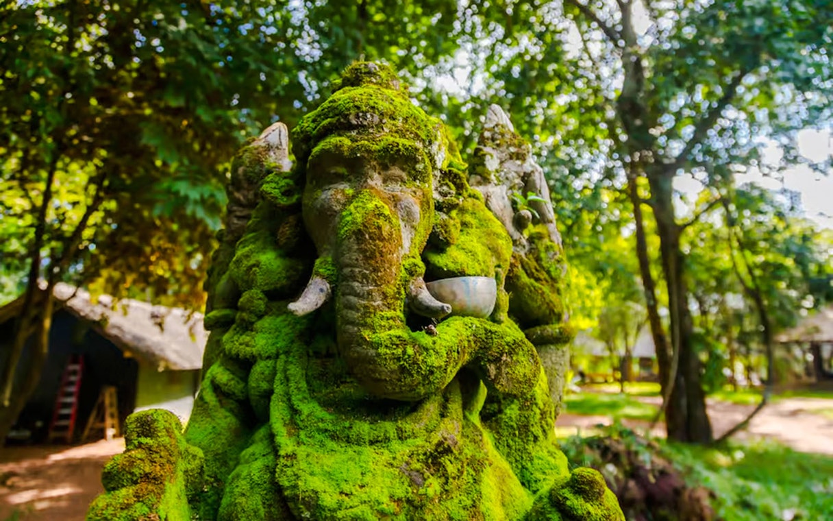 Moss-covered stone statue of Hindu deity Ganesha in a lush forest setting.