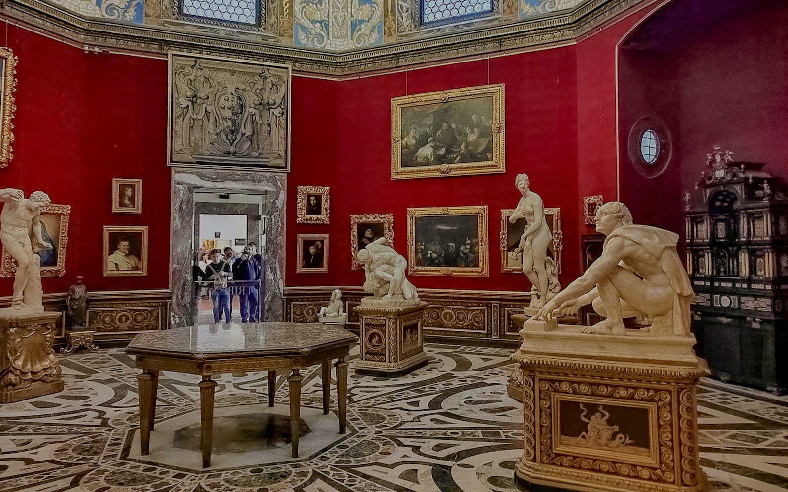 The opulent Medici Gallery immerses visitors in a grand display of sculptures and paintings, enveloped by rich red walls and an ornate ceiling, a testament to the Medici's patronage of the arts