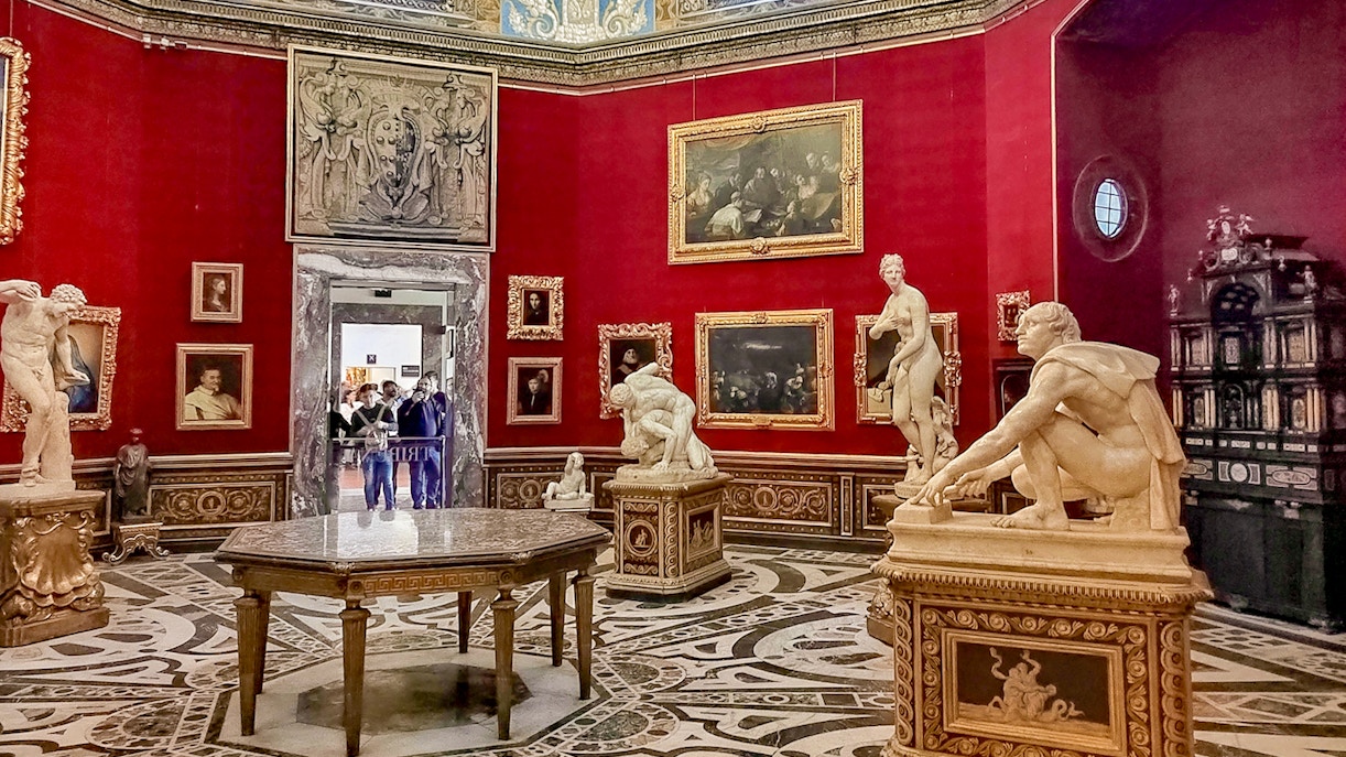 Sala vermelha na Galeria Uffizi, com esculturas renascentistas e luxuosas pinturas emolduradas em ouro, com intrincados desenhos no piso de mármore que aprimoram a experiência artística histórica