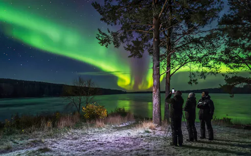 People observing Northern Lights over a lake in Rovaniemi, Finland.