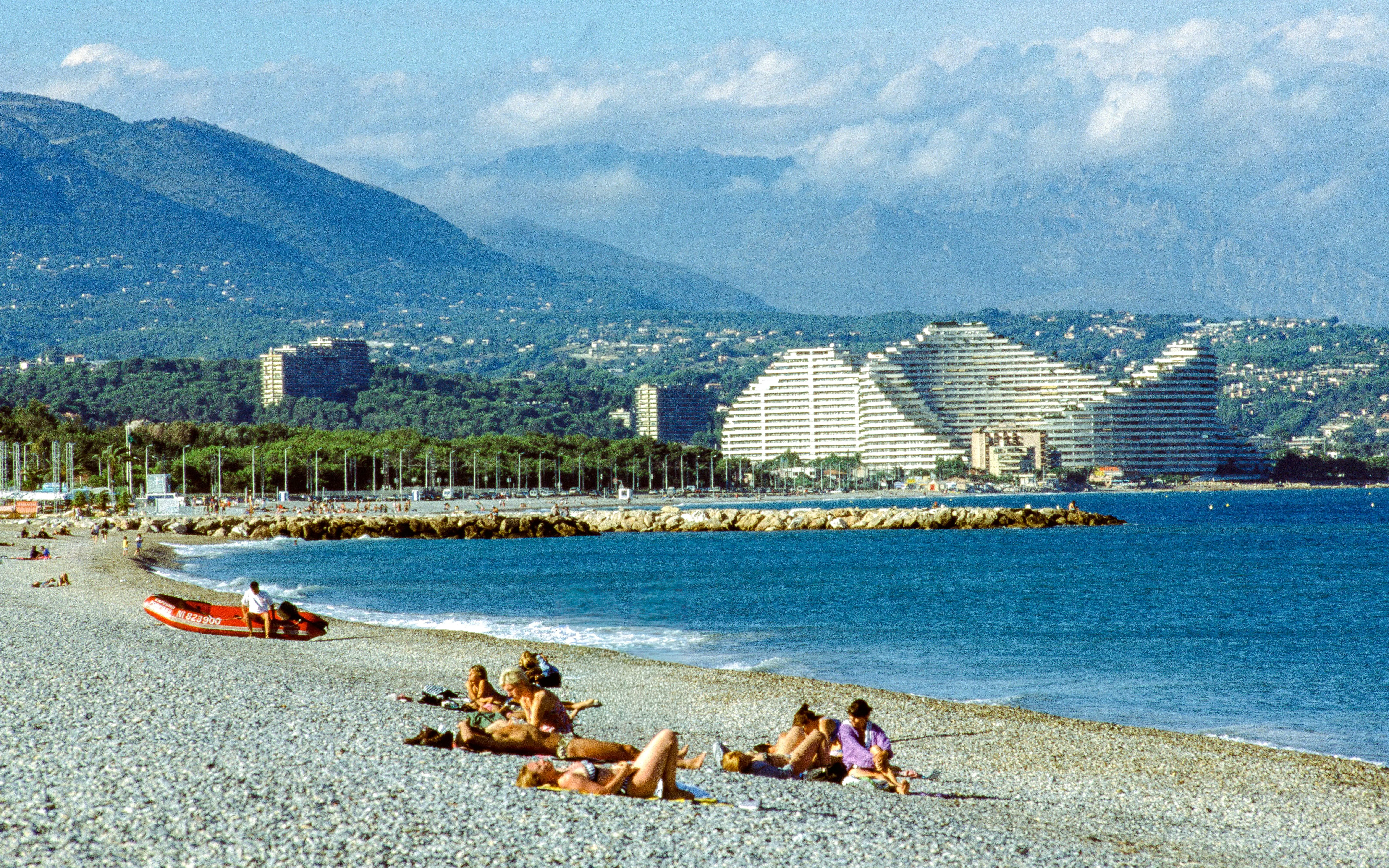 Beachgoers relax on the pebble shore near Marina Very Des Anges with pyramid buildings in the background.