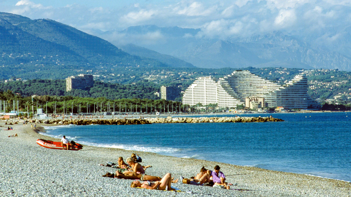 Beachgoers relax on the pebble shore near Marina Very Des Anges with pyramid buildings in the background.