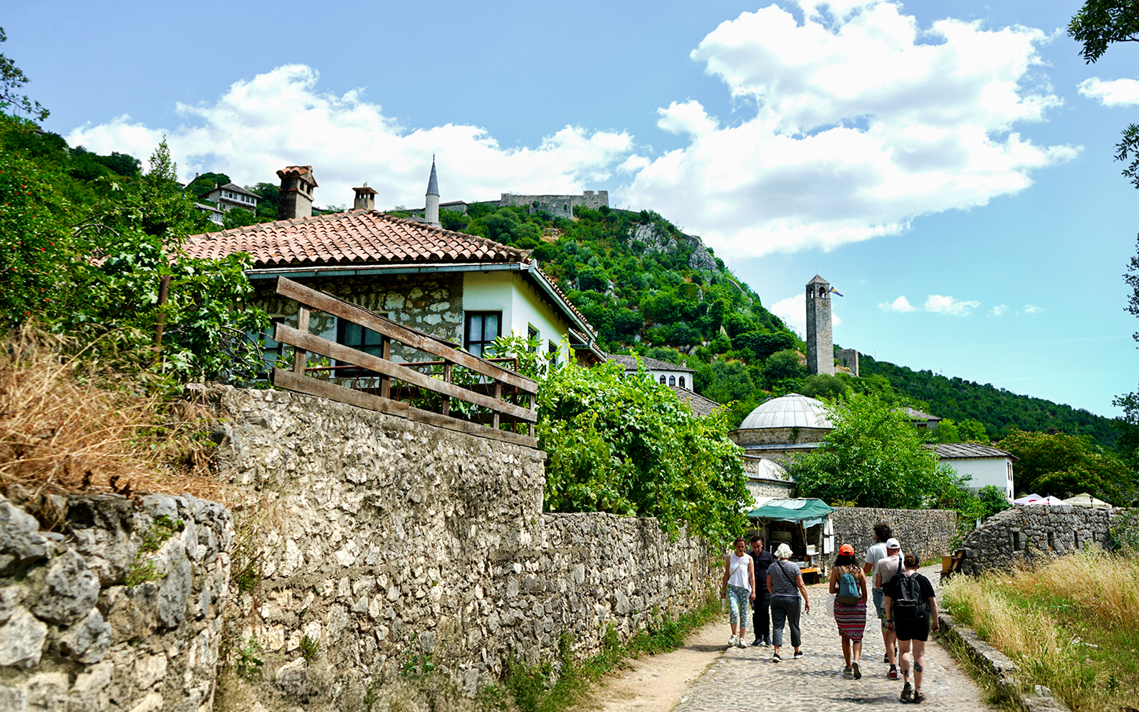 Tourists walking through historic Mostar with stone buildings and a hilltop fortress.