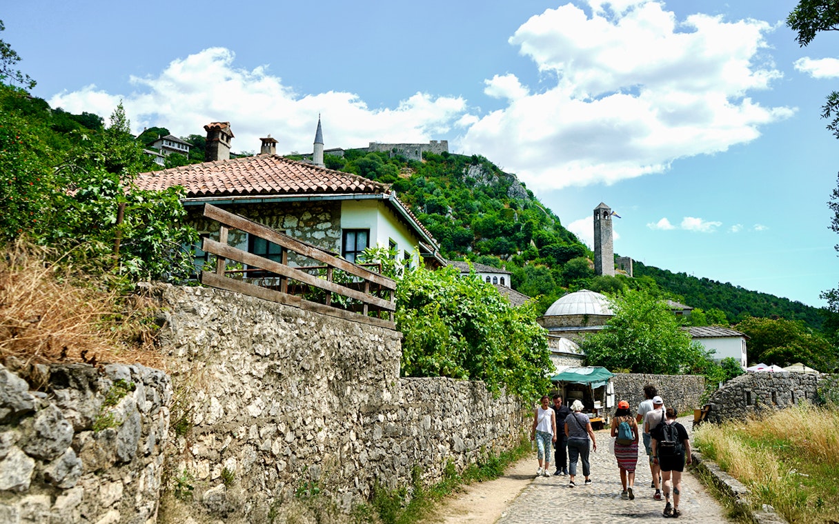 Tourists walking through historic Mostar with stone buildings and a hilltop fortress.
