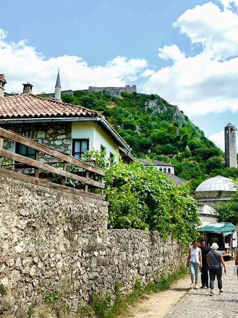 Tourists walking through historic Mostar with stone buildings and a hilltop fortress.
