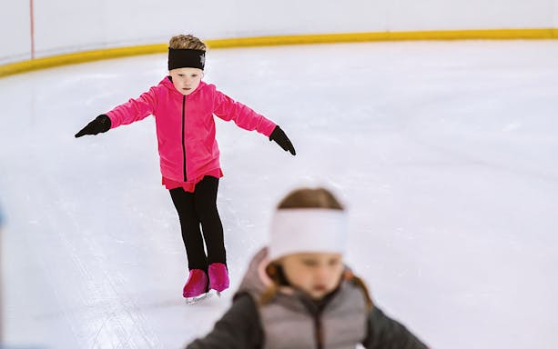 Little girl in pink jacket learning to ice skate on rink.